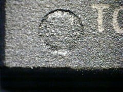 Zoomed in picture of a grey surface of a part with an indent and markings: a white T and part of an O are also visible. The indent is filled up, but not completely.
